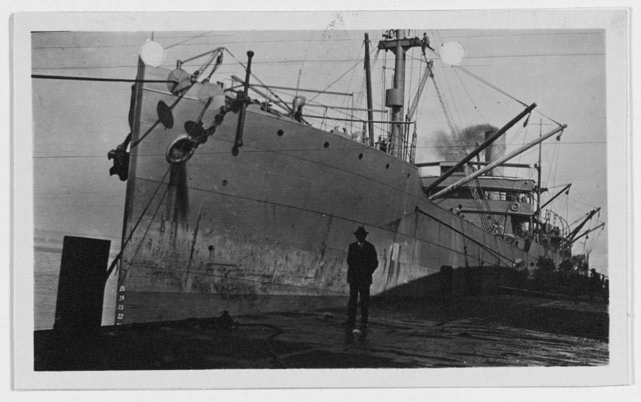 A large single-funnel steam freighter moored alongside a dock, photographed from the pier. A man in a dark suit and hat stands on the dock in the foreground, dwarfed by the hull.