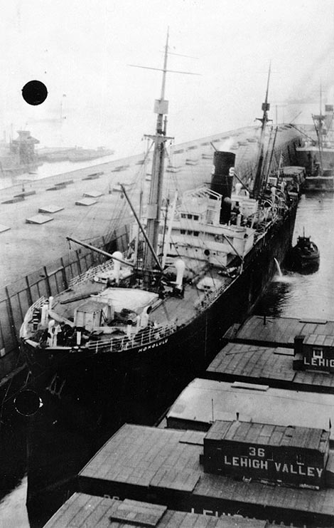 A steam freighter moored in a narrow basin, viewed from above and forward, with the name HONOLULU painted on her bow and Lehigh Valley barges alongside.