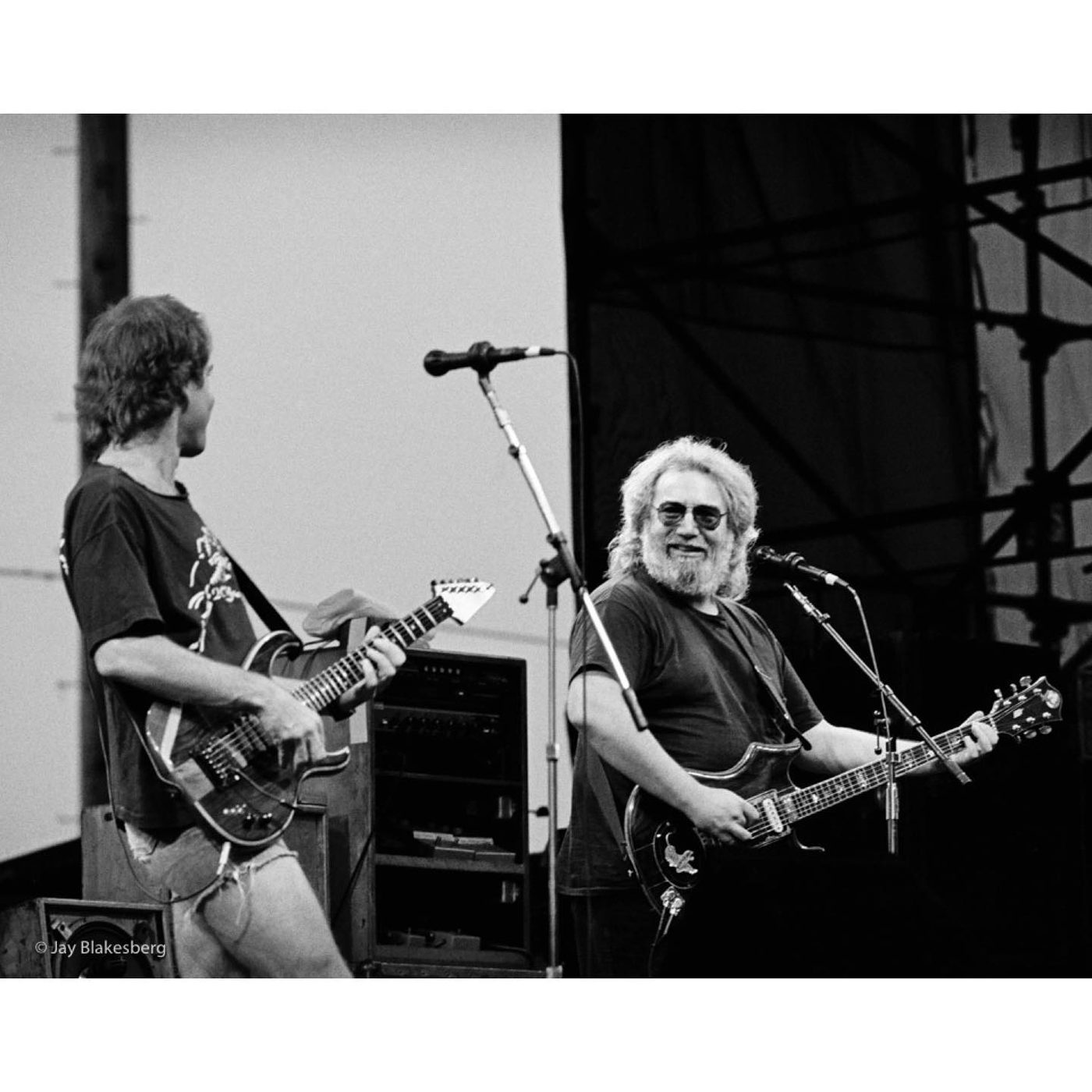 Black and white photograph taken from the side of the stage. Phil Lesh stands at left in cut-off shorts playing bass, facing away. Jerry Garcia stands at right in a dark t-shirt behind a microphone, white hair and beard, round glasses, smiling slightly, playing an electric guitar. Stage scaffolding and a rack of equipment between them.