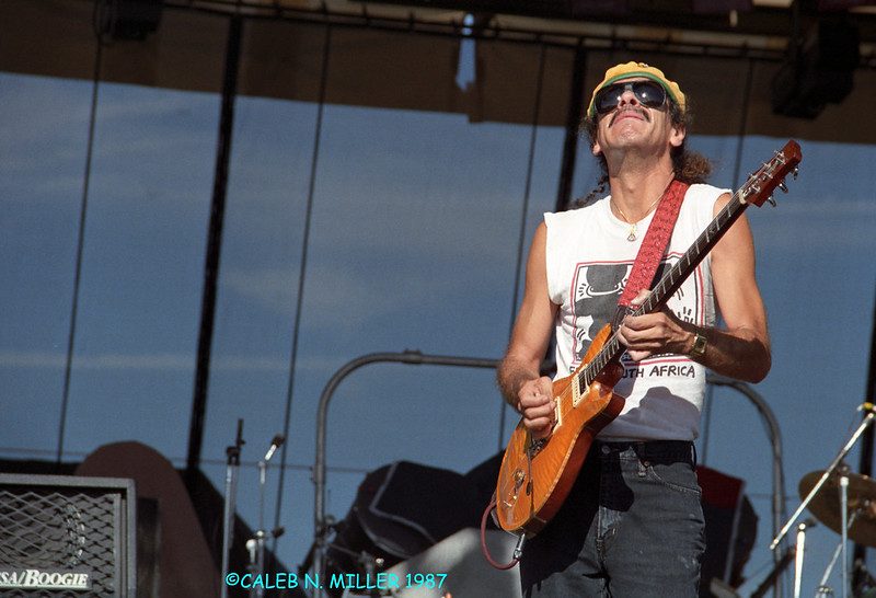 Carlos Santana on stage in daylight, head tilted back and eyes closed behind dark sunglasses, wearing a yellow cap and a white sleeveless shirt, playing a translucent amber electric guitar with a red strap. Blue sky and stage rigging behind him.