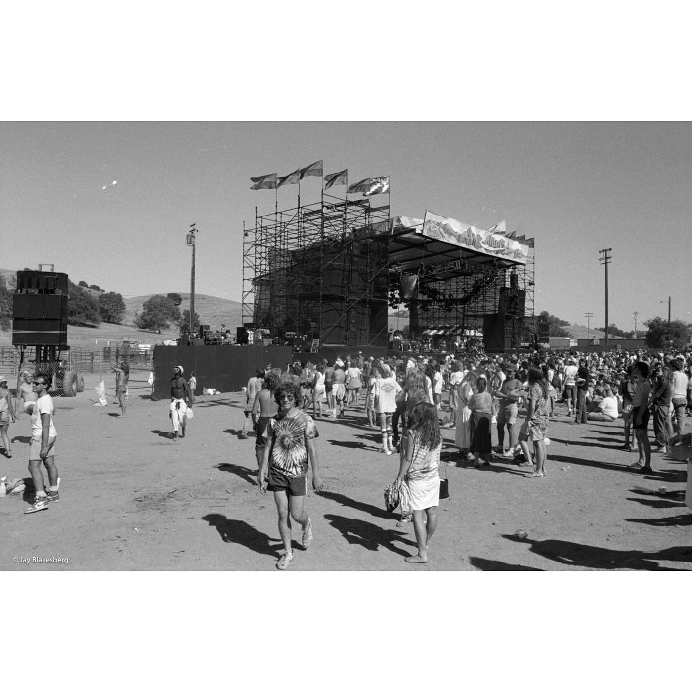 Black and white photograph of a dusty fairground in bright sun. A large scaffolded stage stands at center with flags flying from its towers, one bearing a Steal Your Face design. Several hundred people in tie-dye and shorts stand and sit on bare dirt in front of it, with dry golden hills behind.