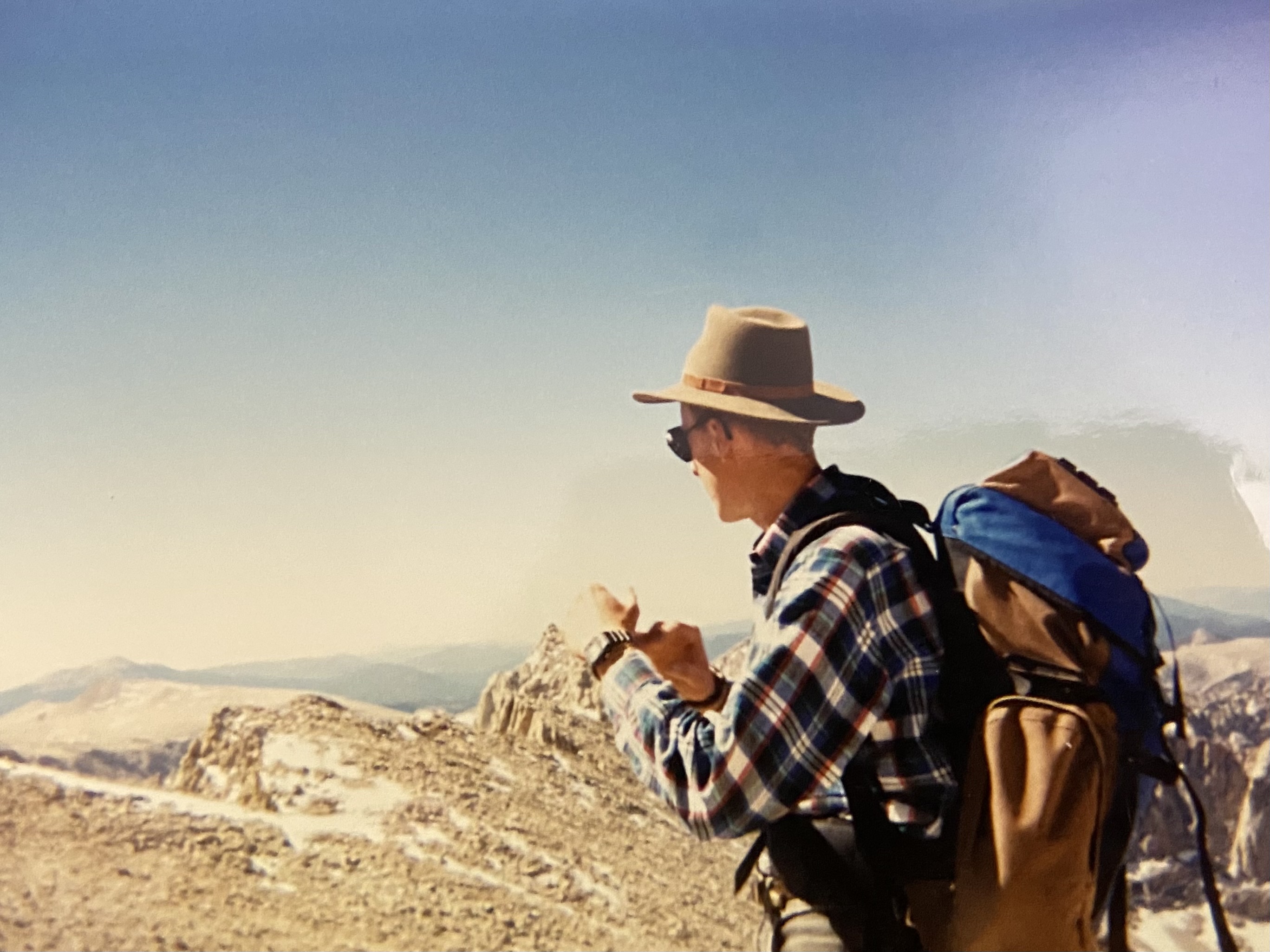 Ben, the Austrian, in a plaid flannel shirt and a wide-brimmed felt hat, external-frame pack on, checking his watch, high peaks behind him.