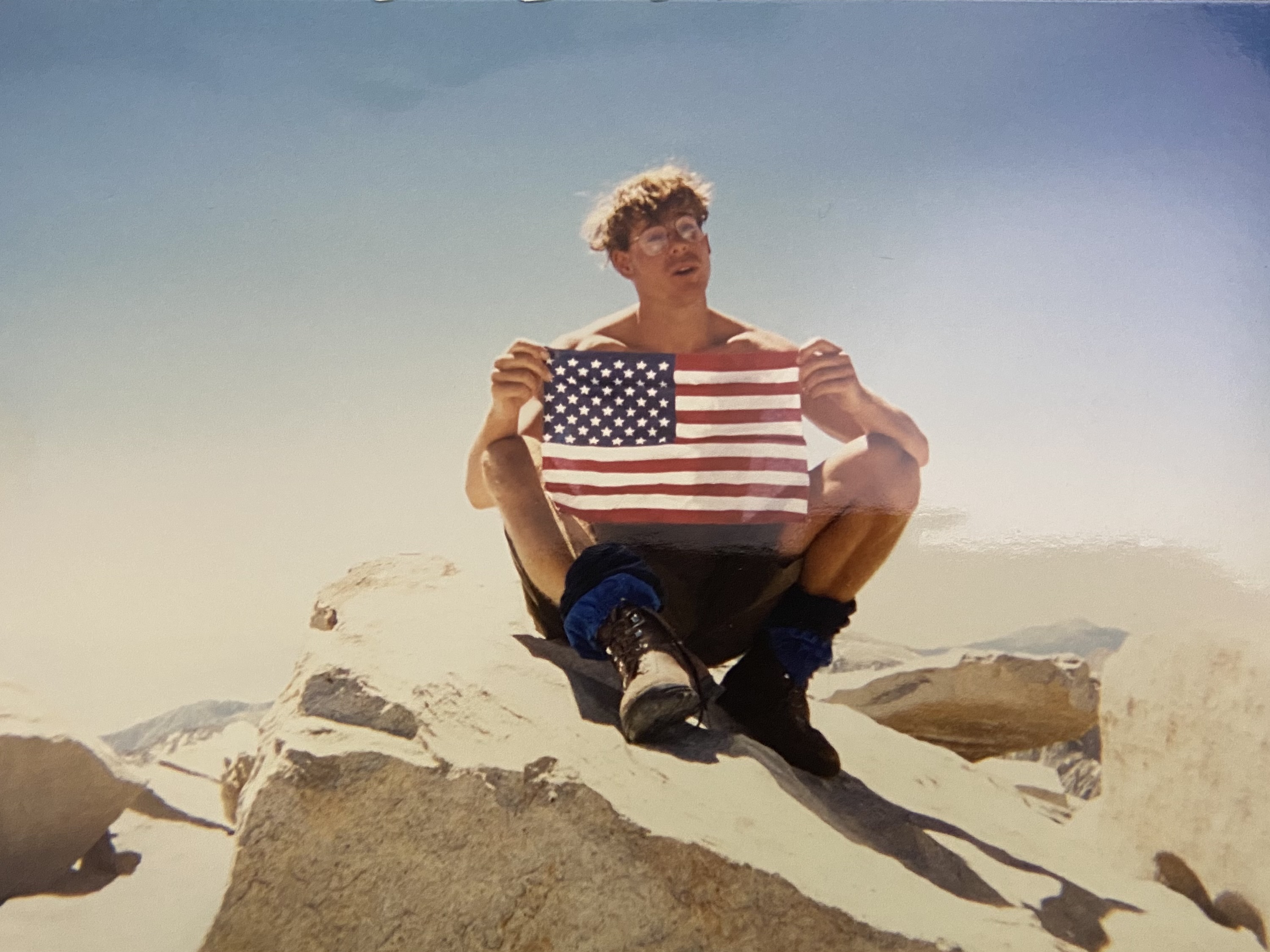 Brooks sitting cross-legged on the summit rocks, holding a small American flag open in front of him, glasses on, hair wild, squinting into the light.