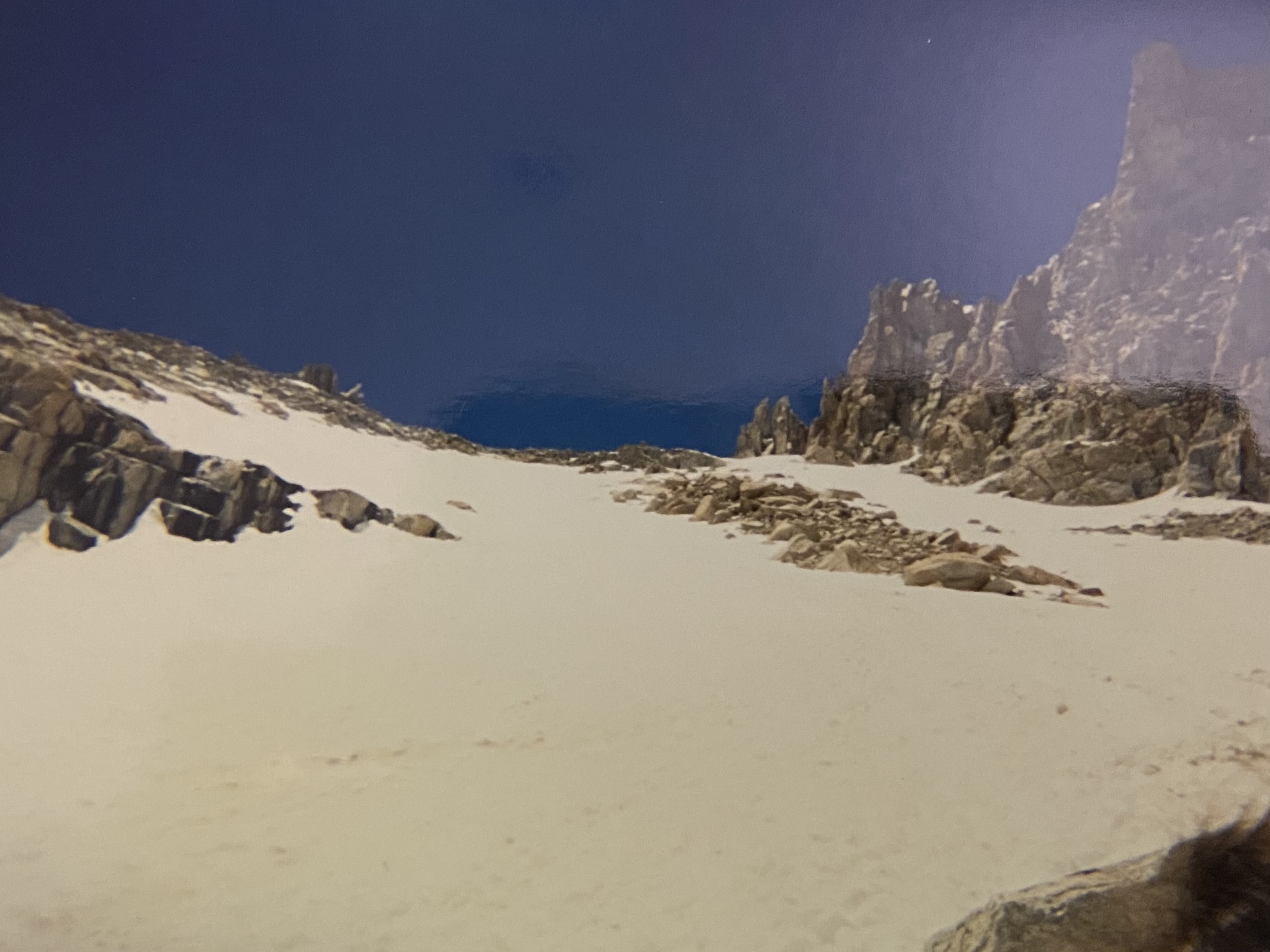 The snow-filled cirque below Trail Crest: an unbroken white slope running down and out of frame, dark granite ribs and pinnacles rising on the right, deep blue sky above.
