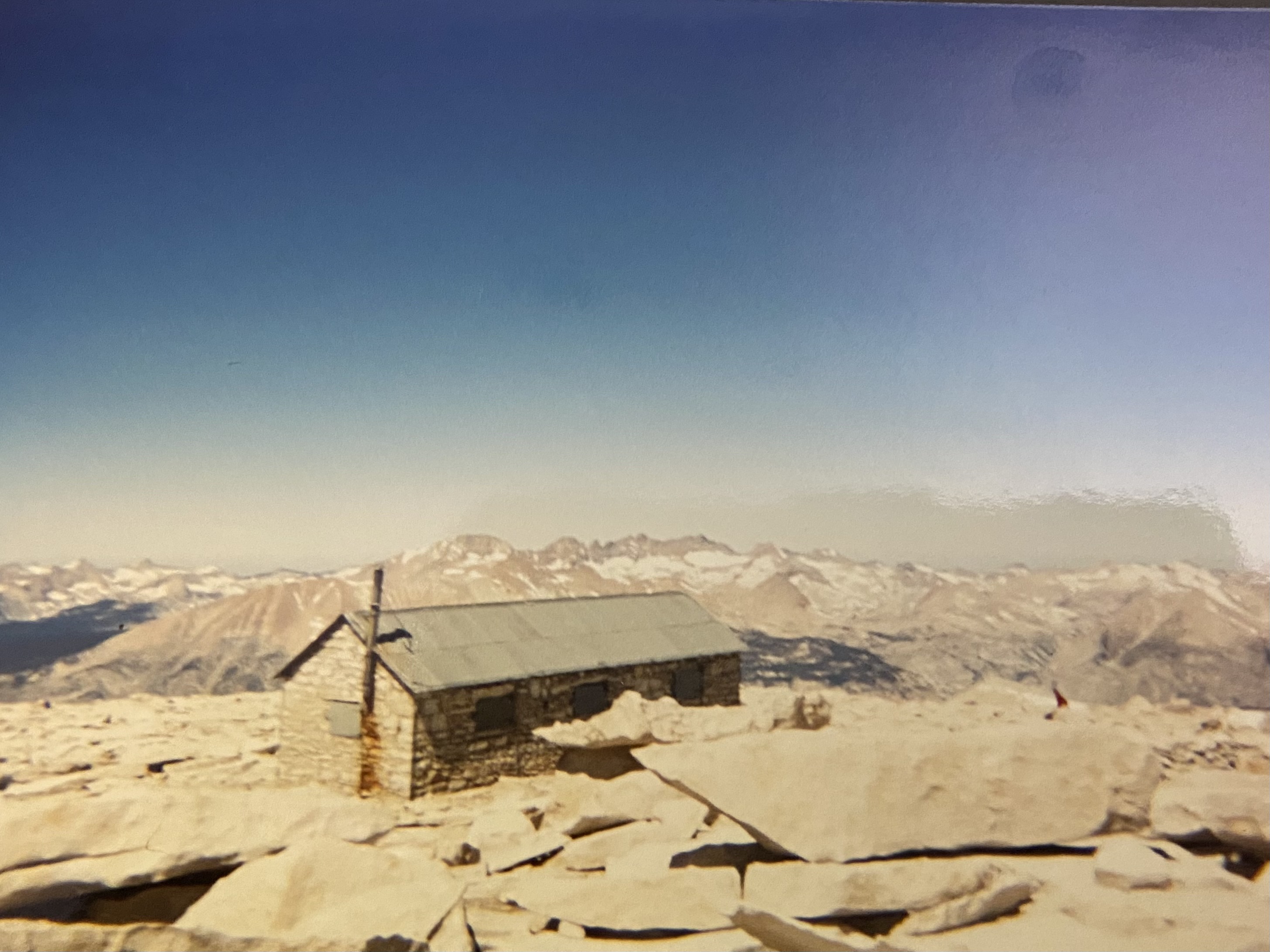 The stone Smithsonian shelter on the summit of Mt. Whitney, metal roof, snow patches around it, the Great Western Divide running off behind.