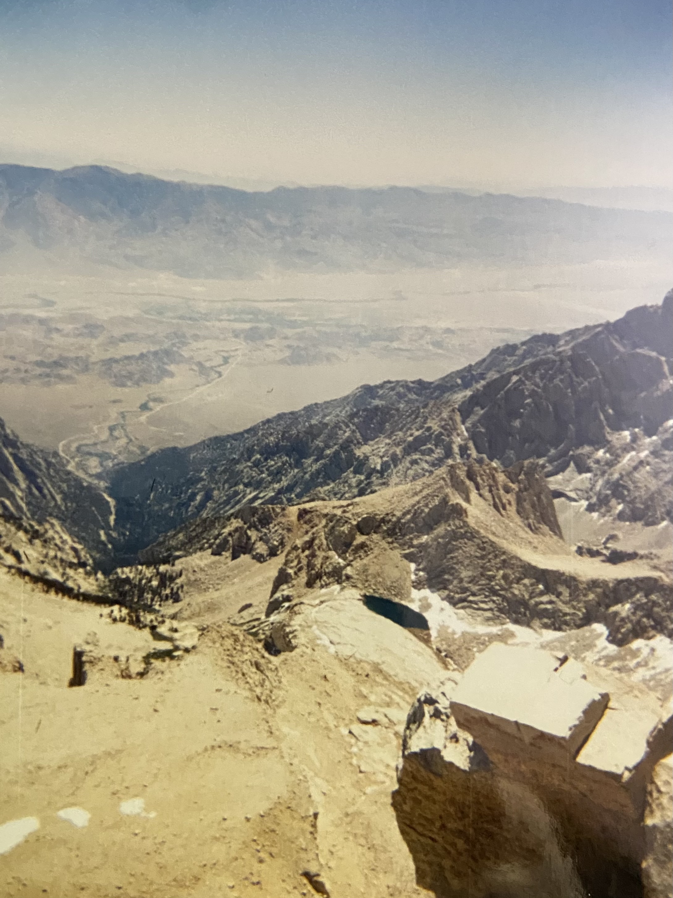 Looking east from the summit of Mt. Whitney down the east face: the Owens Valley eleven thousand feet below, Owens Lake bed hazy in the distance, a small tarn and snow patches in the cirque directly under the drop.