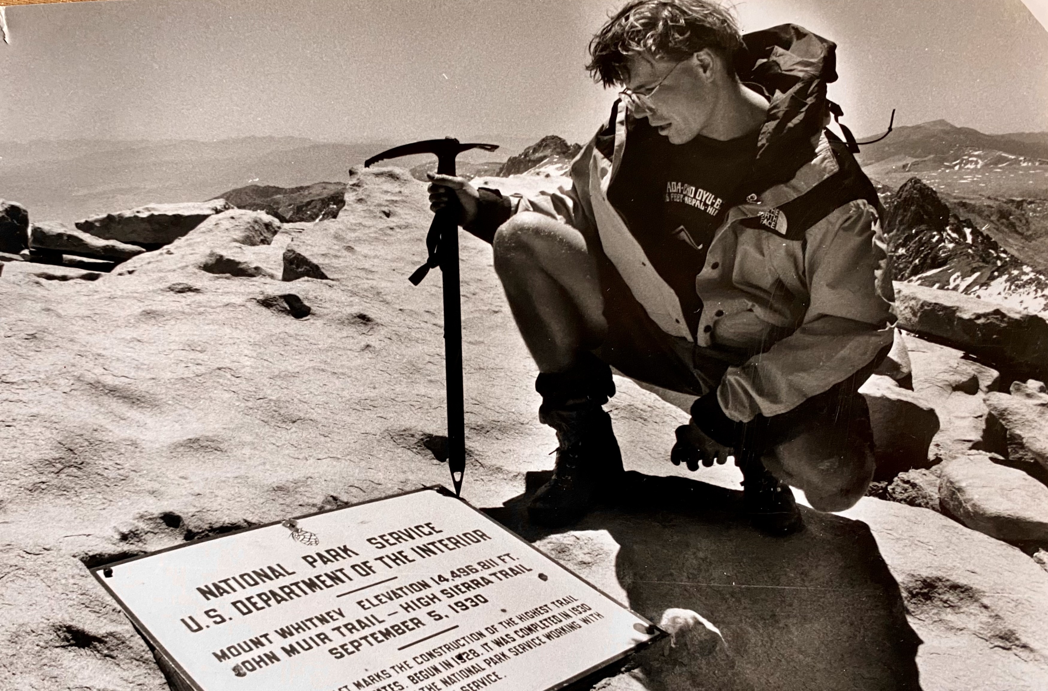 Black and white photograph of Brooks crouched on the summit of Mt. Whitney reading the elevation marker, ice axe planted beside him