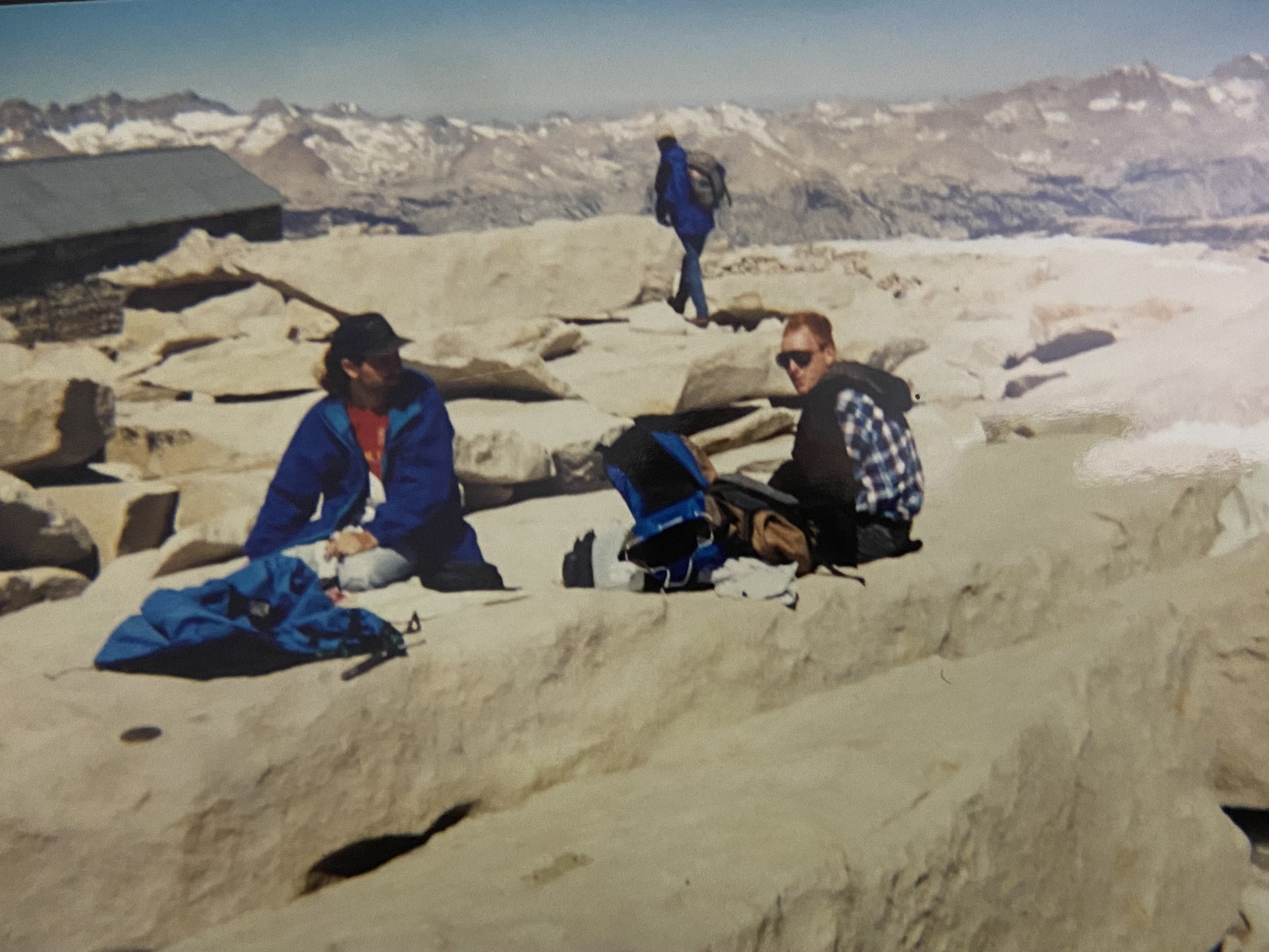 Zack Hurley and Ben sitting on the summit slabs beside the stone hut, eating, with packs and jackets spread around them, a third figure walking away in the background, snowy ranges behind.