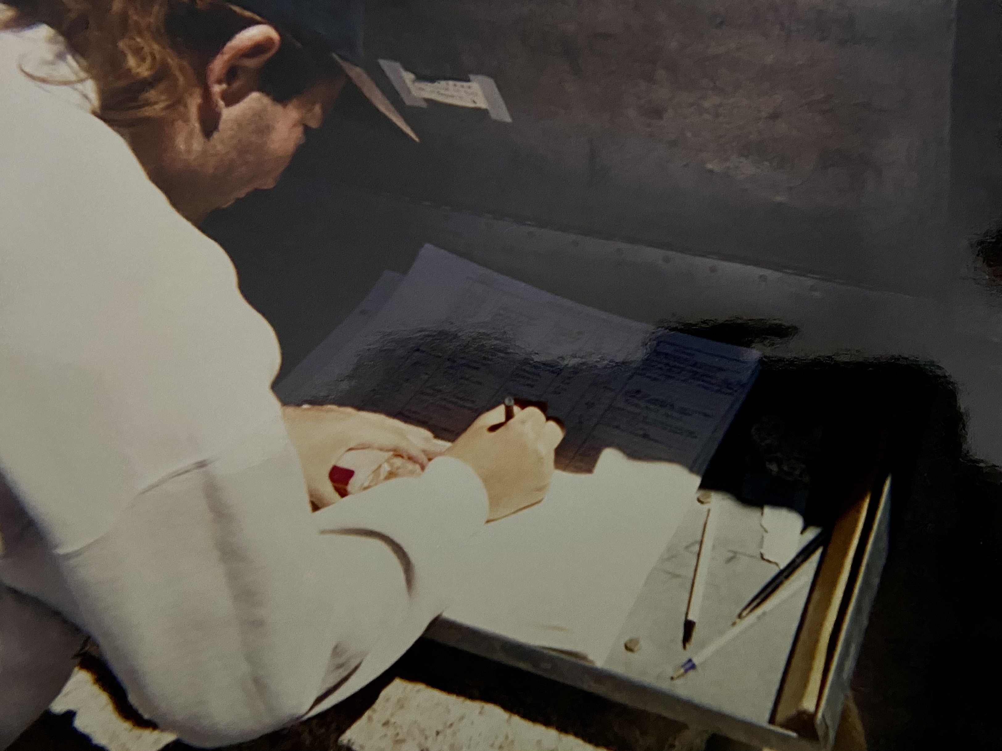 Signing the summit register inside the shelter: a figure in a white sweatshirt and ballcap bent over the open logbook, pens and papers on the metal box.