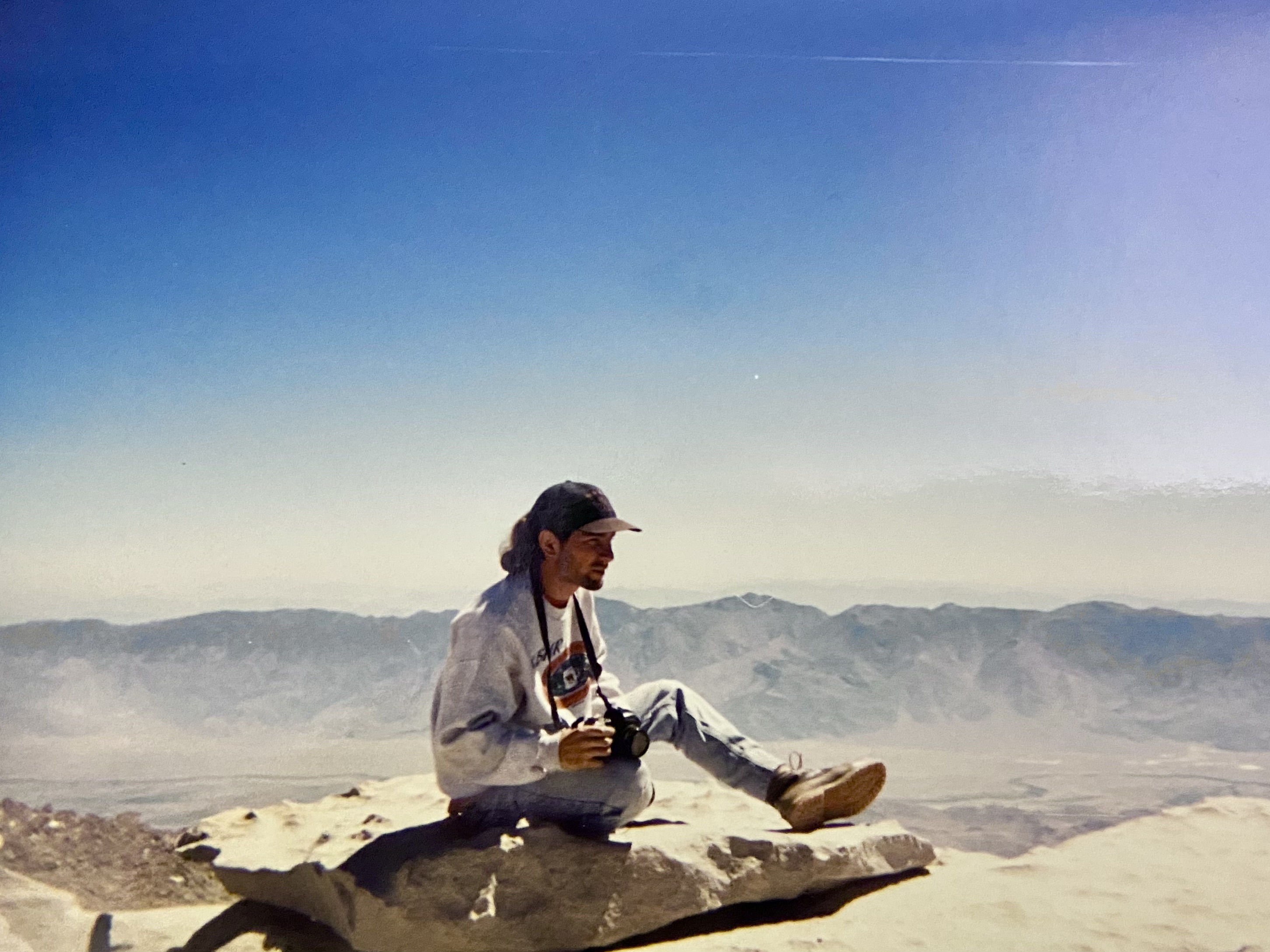 Zack Hurley sitting on a flat slab of granite at the summit edge in a grey KBHR sweatshirt and ballcap, Nikon in his hands on its strap, the Owens Valley and the Inyo Mountains hazing out far below and behind him.