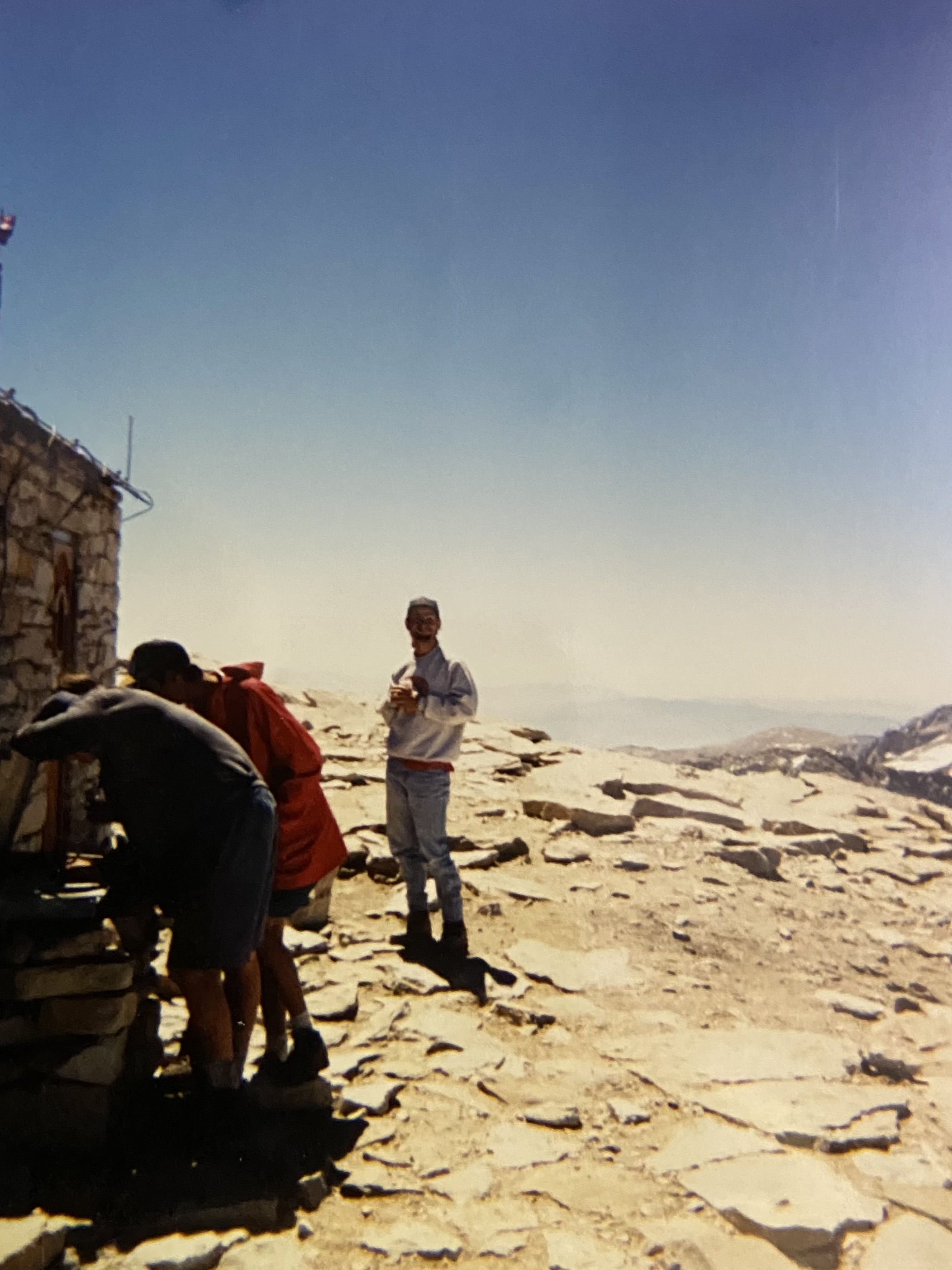 Zack Hurley standing on the summit in a grey sweatshirt and ballcap, waiting his turn to sign the register, ranges receding behind him.