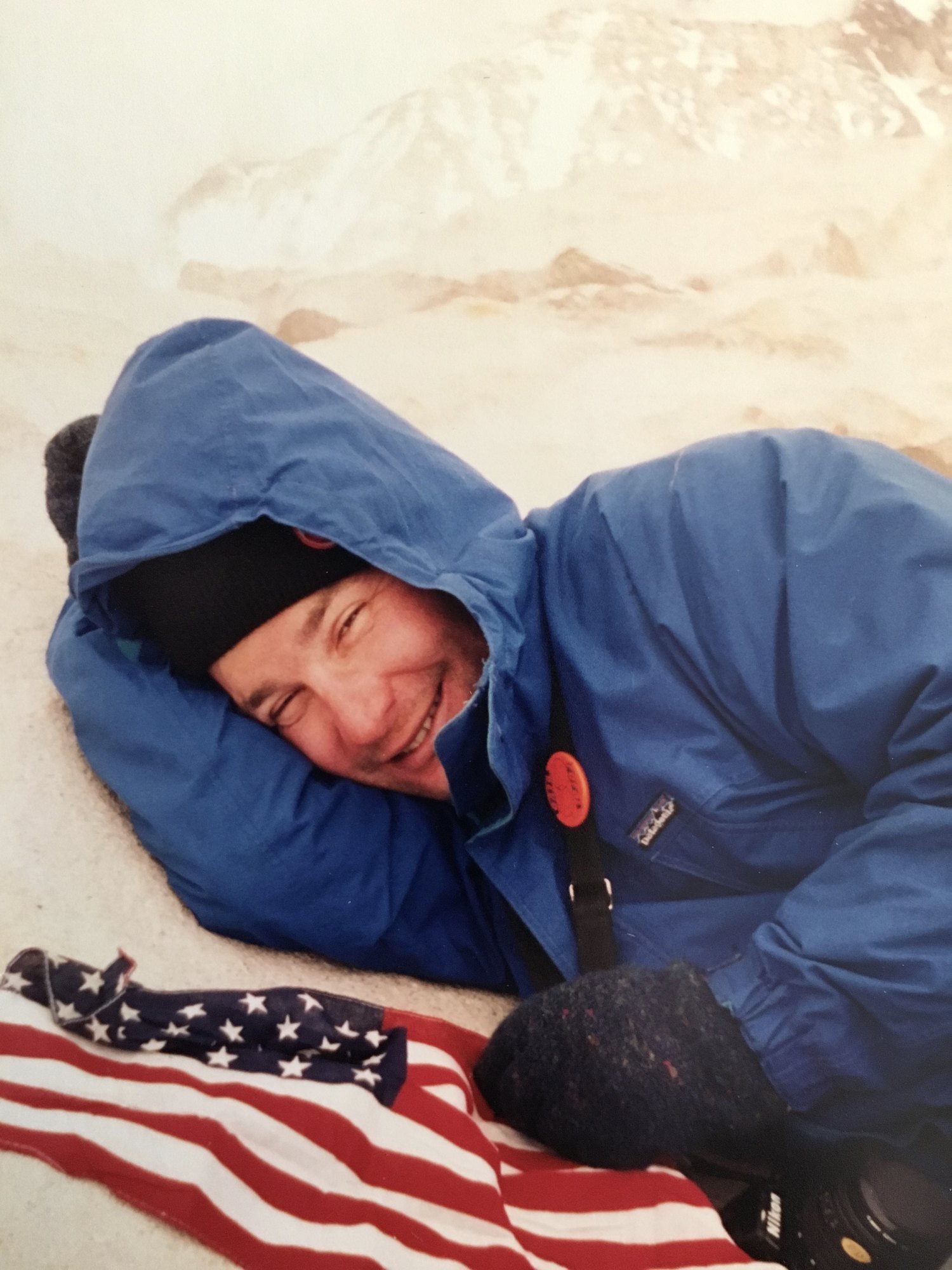 Zack Hurley lying in the snow near the summit, grinning, an American flag spread out beside him and his Nikon at his side.